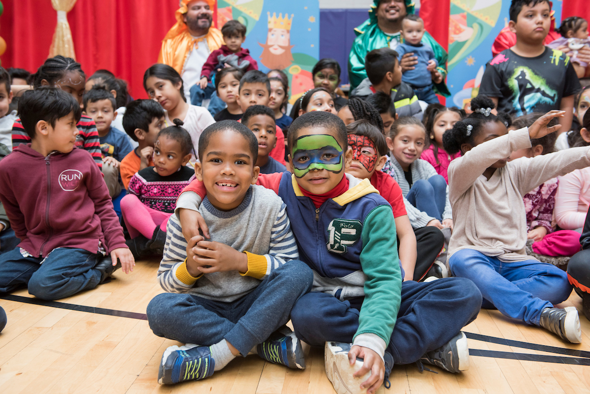 About 25 small children sit on a wooden gymnasium floor smiling for a photo in front of several men in shiny robes dressed as the Three Kings. Several children have their faces painted in festive colors.