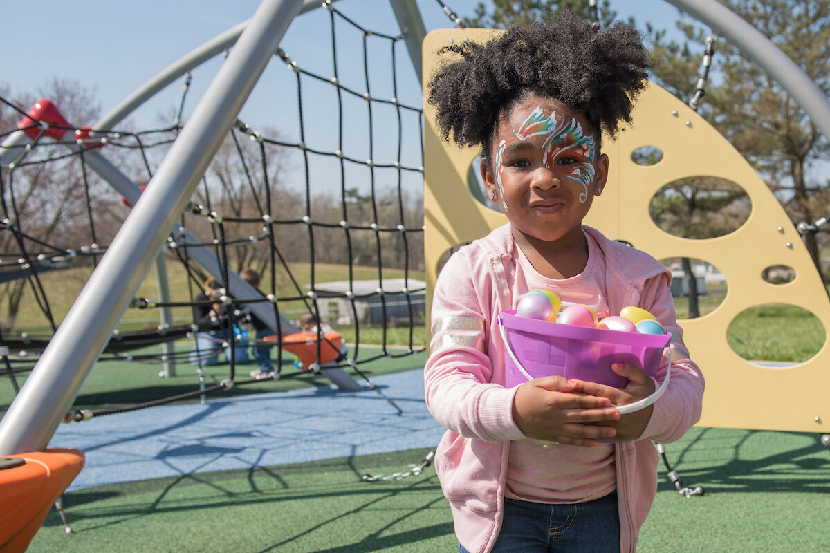 Outside on a sunny playground, a small girl stands holding a plastic bucket full of plastic Easter eggs. Her face is painted with rainbow butterflies.