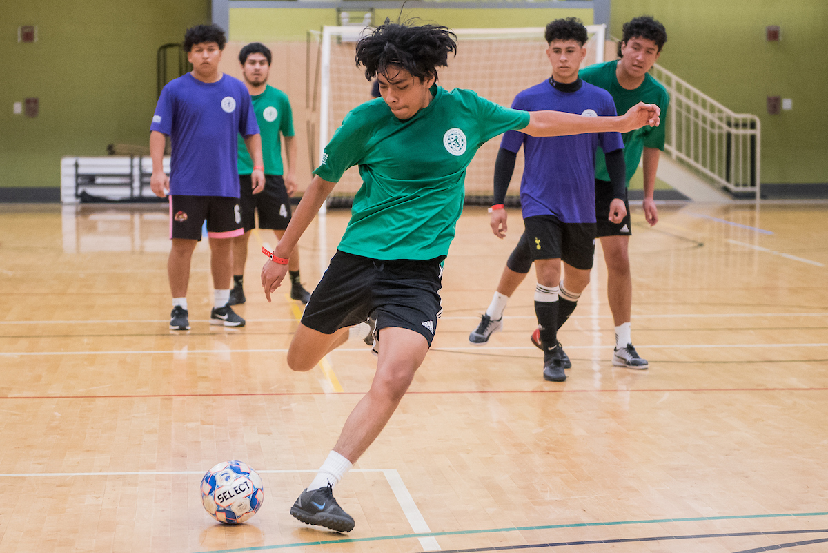 A game of futsal is played in a gymnasium between players wearing green shirts and players wearing purple shirts. A player in a green shirt prepares to kick the ball while others look on.