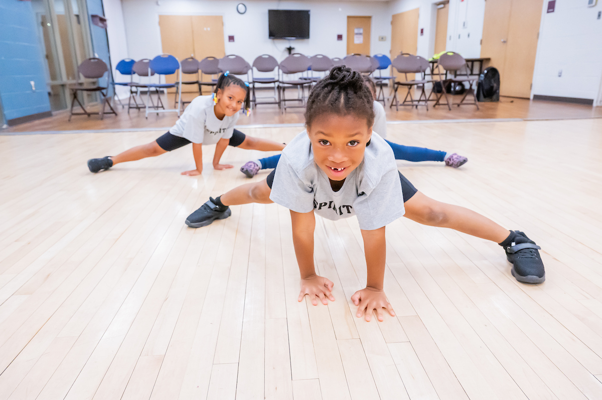 In a dance studio with a hardwood floor, three kids demonstrate the splits while grinning at the camera. Behind them in the room, rows of chairs are set up facing the dance floor.
