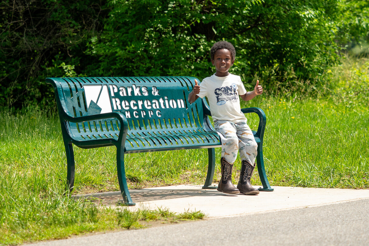 A small child sits on the far end of a green metal park bench with the words Parks & Recreation on it. The child is smiling and giving two thumbs up to the photographer. The bench sits on a concrete pad adjoining an asphalt walking path. Grass surrounds the concrete pad, with green brush and trees in the background.