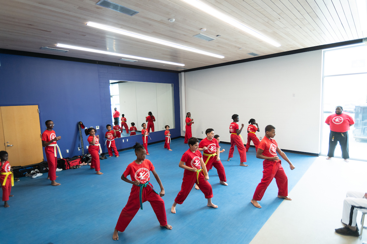 In a large studio with a mirror on one wall, 14 youth in matching red martial arts uniforms practice martial arts. They wear different colored belts, some are yellow, some are white, and a few are blue. Their instructor stands off to the right side against a window, with his arms folded behind his back, watching the class.