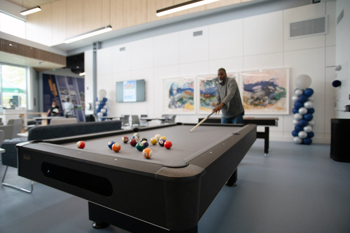 In a large lounge-type room with three paintings hanging on the far wall, a man plays pool at a pool table covered in gray felt. Behind the pool table are cafe-style tables and chairs. There are two balloon decorations in the room, made of silver and blue balloons.