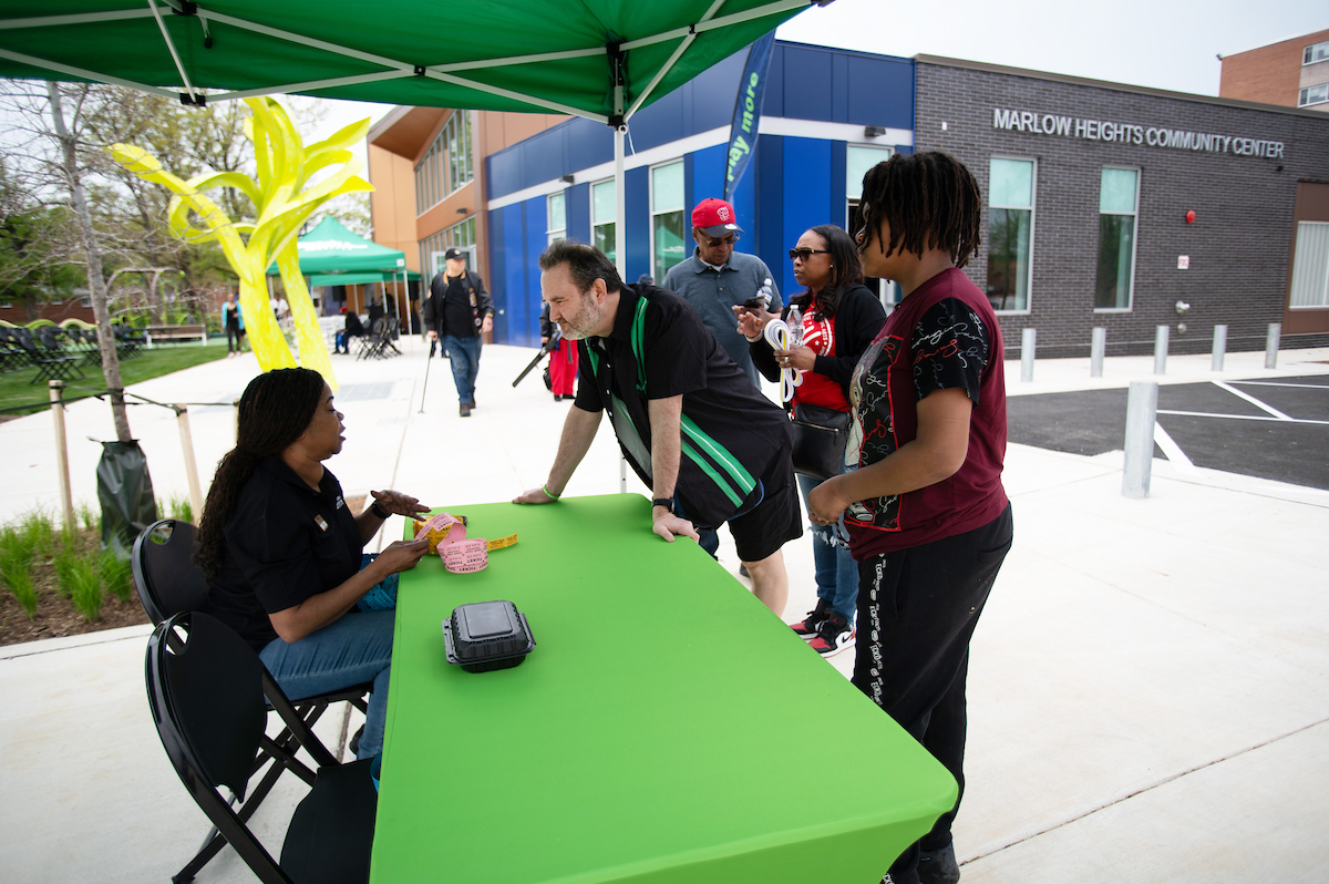 On a concrete patio next to a modern brick and glass building, a woman sits at a green check-in table under a green event tent, greeting customers. Letters on the building in the background read MARLOW HEIGHTS COMMUNITY CENTER. Behind the table to the left is a large, neon yellow sculpture and more green event tents.