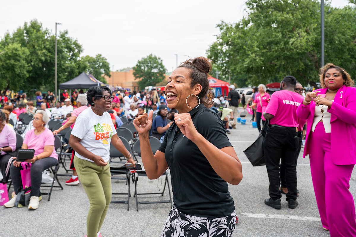 Several people dance in a parking lot that has been converted to a concert venue, with folding chairs for the audience. Several event tents are set up in the background.