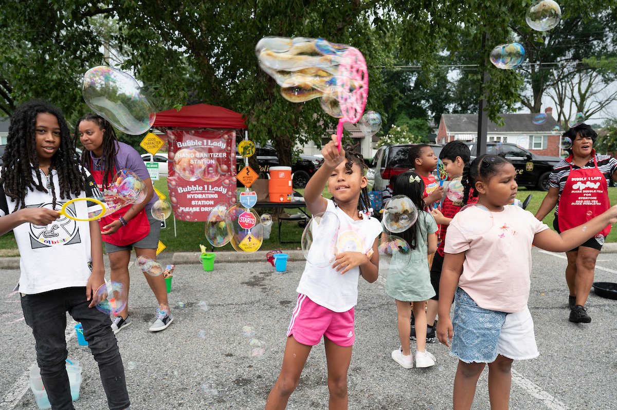 Half a dozen children use bubble wands of various sizes to create soap bubbles in an asphalt parking lot, at a station with a sign that reads Bubble Lady. Two women in red Bubble Lady aprons are assisting.