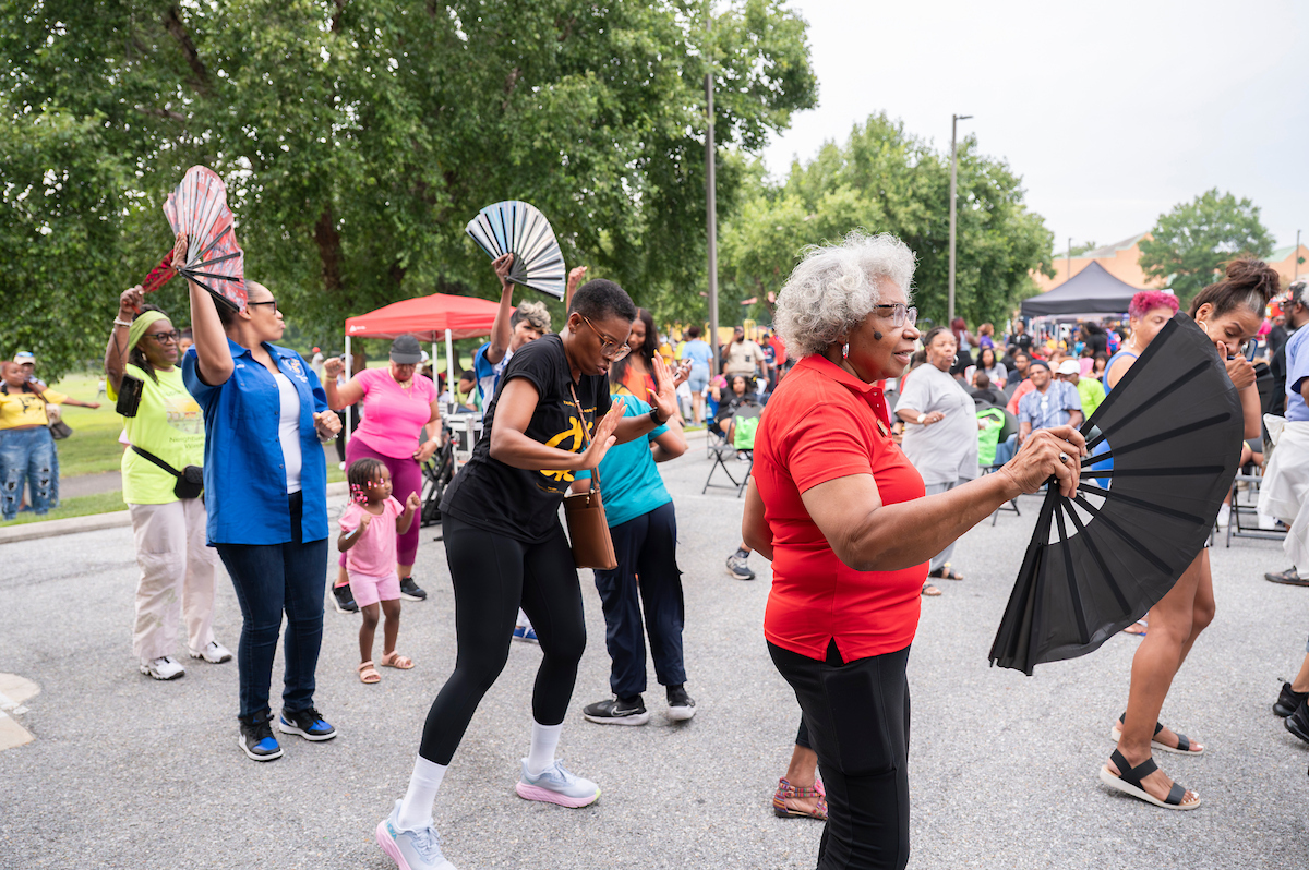 A group of women with hand-held fans dance together in an asphalt parking lot. Behind them are rows of folding chairs filled with people, and several event tents with people crowding around them.