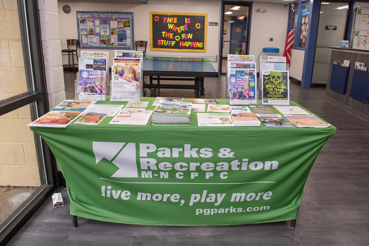 Inside a lobby area, a table with a green Parks and Rec tablecloth contains dozens of neat stacks of flyers, program guides, and literature. In the room behind this is a ping-pong table and a bulletin board; to the right, the edge of a reception desk is just visible.