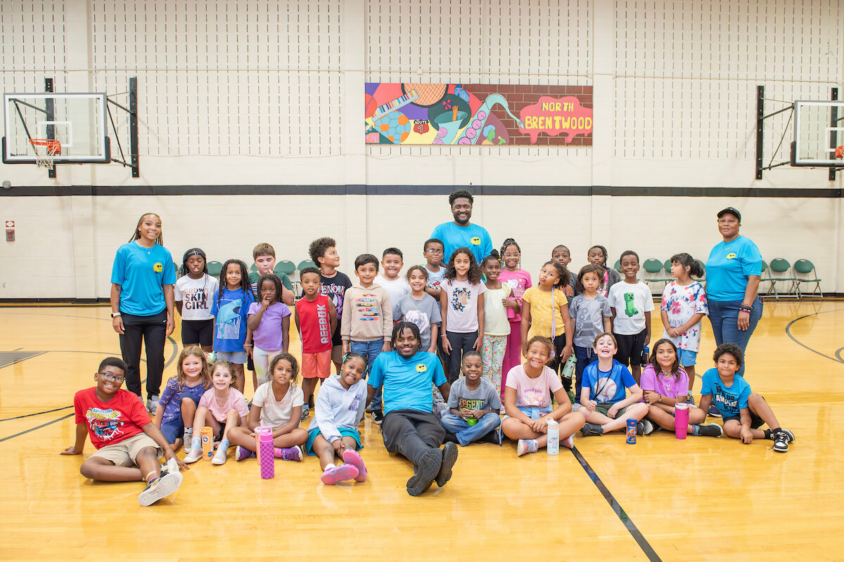 About 30 kids pose for group photo with four summer camp counselors in a large gymnasium. A colorful mural banner on the wall of the gym reads NORTH BRENTWOOD and incorporates motifs of musical instruments and sports equipment.