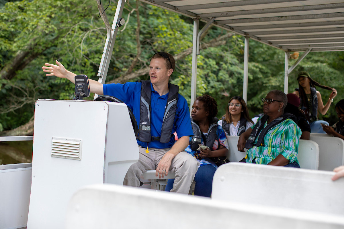 A guide driving a pontoon boat gestures broadly with one arm while giving a boat tour. Behind him are several rows of passengers in low seats, looking off in the direction he is gesturing. They are all wearing inflatable PFDs around their necks. Outside the boat are green trees above brown water.