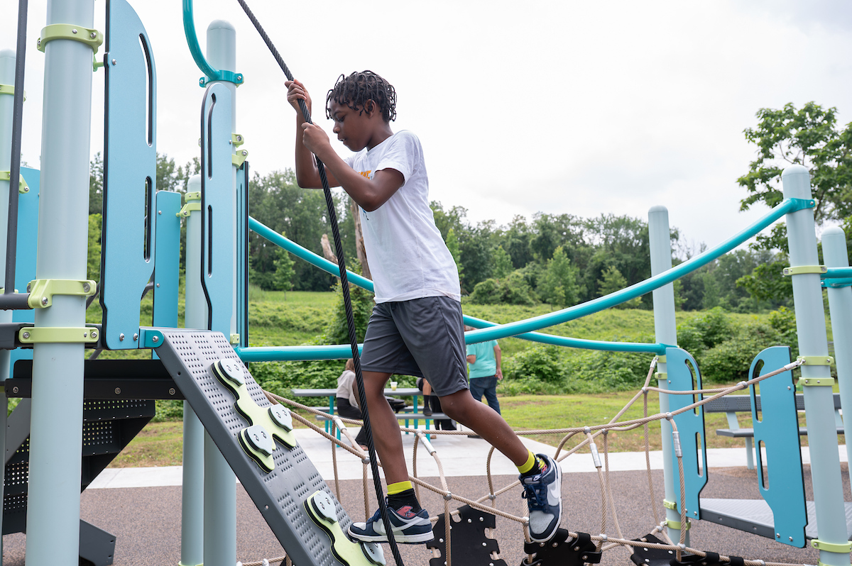 A boy holds onto a rope while climbing up a steep climbing wall feature at an outdoor playground. Adults sit around a picnic table in the background.