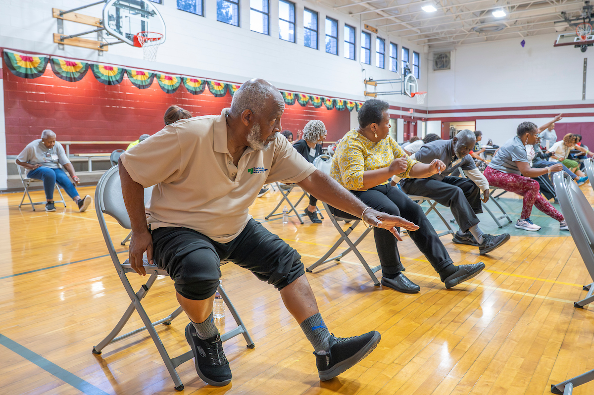 In a gymnasium, seniors sit in rows of folding chairs doing seated strength exercises. They are all extending their left legs and arms while looking at the class instructor.