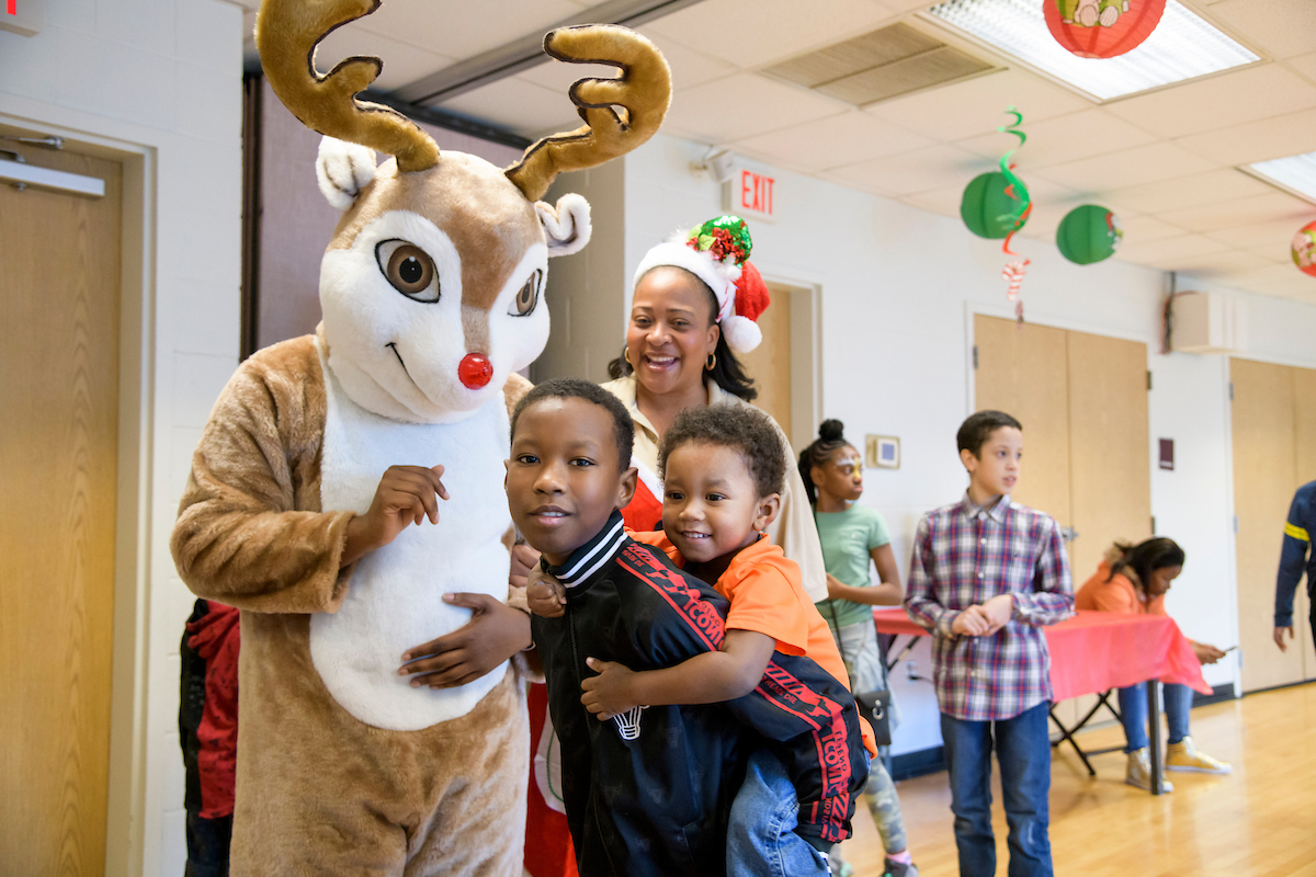 In a room festively decorated for the holidays, a woman in a Santa hat and two boys pose next to someone in a furry Rudolph the Reindeer suit. The larger boy is giving the smaller boy a piggyback ride.