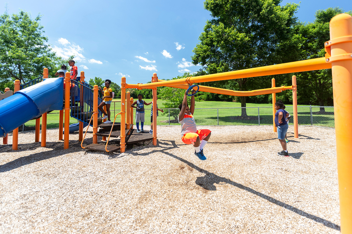 On an outdoor playground, a child hangs from a spinning ring feature attached to a large, blue and gold play structure. The play structure also features slides, platforms, and climbing elements.