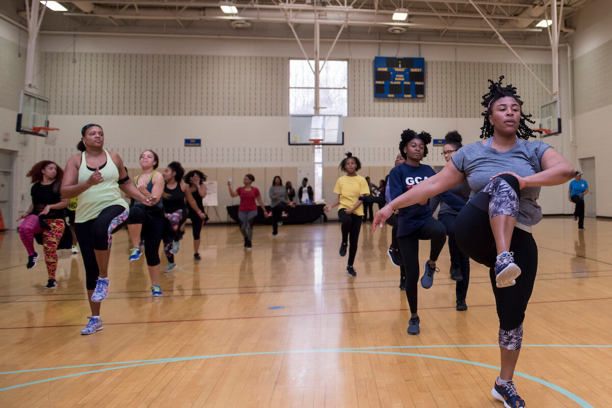 In a gymnasium, several dozen women and teens in athletic clothing exercise in a Zumba class.