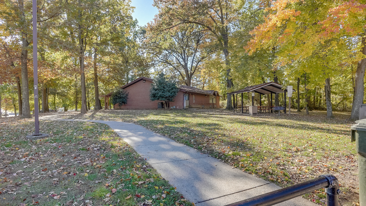 A sidewalk leads through a grassy lawn toward a single-story brick building surrounded by trees. Behind the building is a picnic shelter.