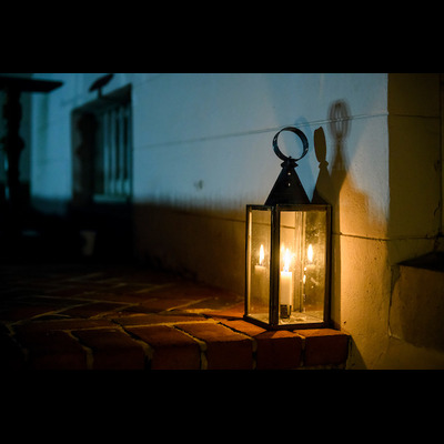 A candle burns inside a glass and metal lantern that is sitting on a brick surface next to a white wall.