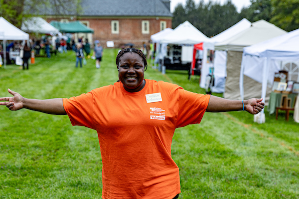 Person standing on a grassy field with arms outstretched, wearing an orange “Volunteer” T‑shirt, with rows of white vendor tents and people walking between them in the background.