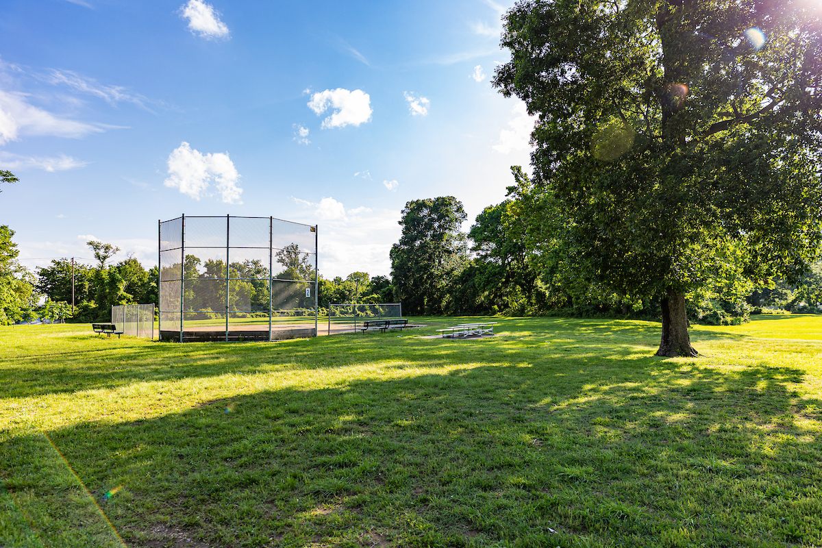 In a grassy field is a small baseball or t-ball diamond with a chain-link backstop behind home plate. There is a large oak tree behind the bleachers and other trees in the far distance.
