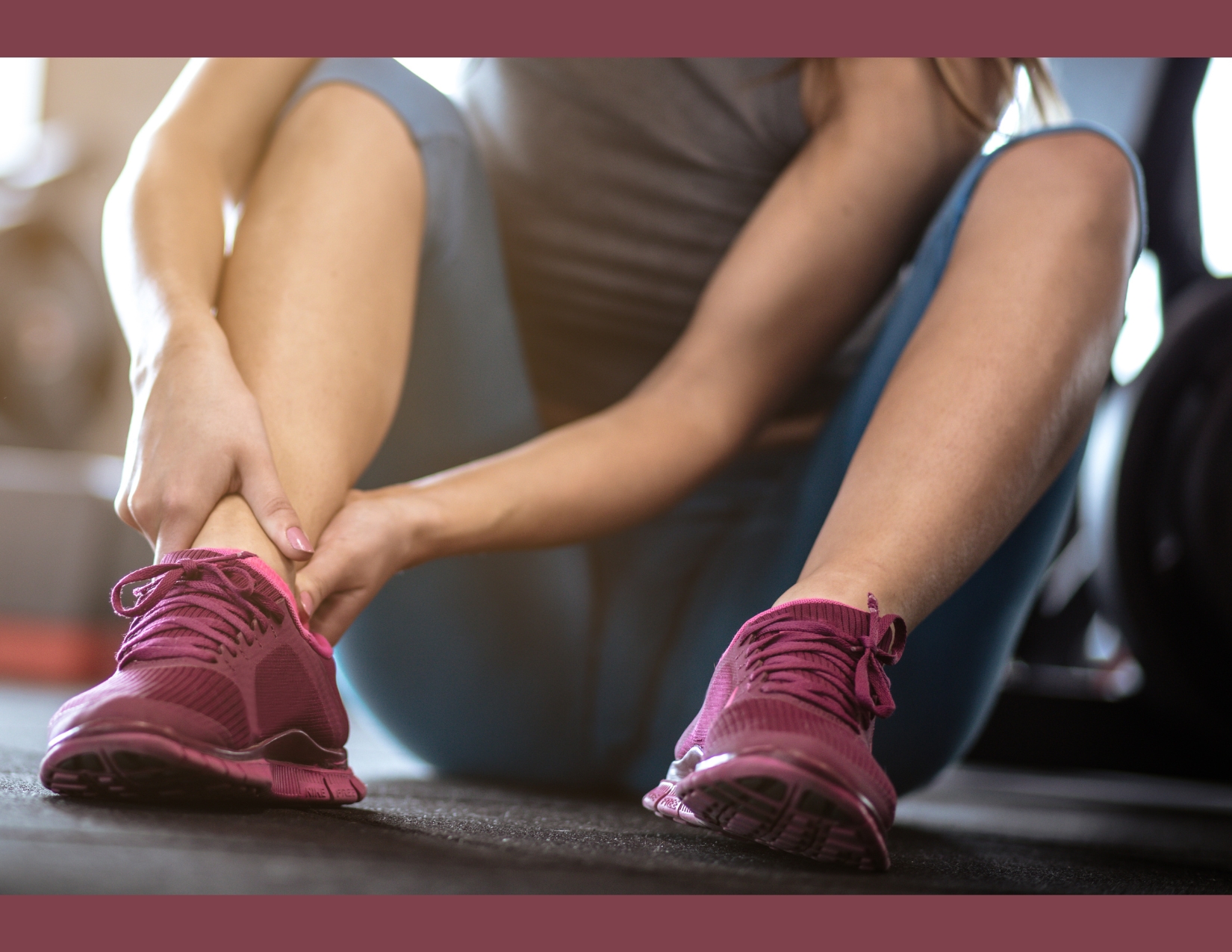 Close-up view of a woman sitting on the floor wearing exercise clothes and maroon sneakers, grabbing one ankle with both hands.