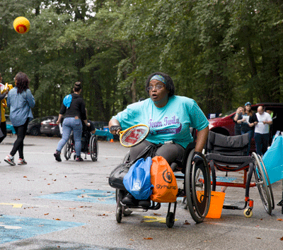 Outside in a parking lot, a woman in a wheelchair holds a tennis racquet and prepares to hit a ball that is sailing toward her. Behind her are other people and several wheelchairs.