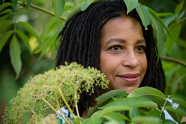 A woman standing in dense foliage. Her head can be seen peeking through leaves, and she's holding herbs in her hand.