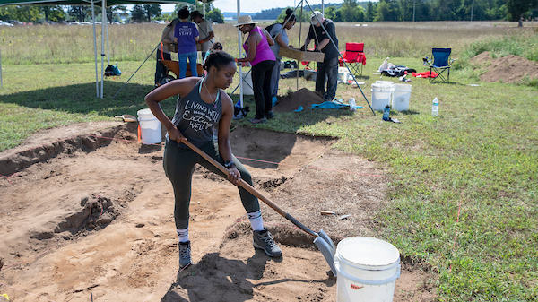 A person standing and digging in a field during the day holding a shovel next to a white bucket.