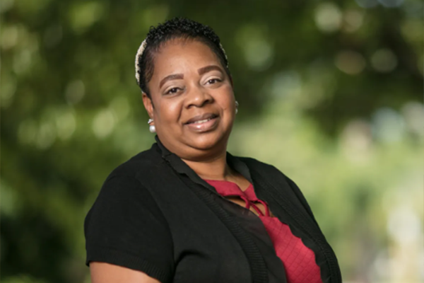 An African American woman with a black and red shirt. She is standing in front of lush green trees in the background