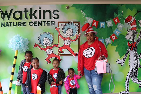 An adult standing with their four children. The children are each wearing a shirt that says "Thing 1, 2, 3 and 4." The adult is wearing a shirt that says "Teacher of all things." They are standing against a Dr. Seuss decorated background with the Watkins Nature Center sign in the corner.