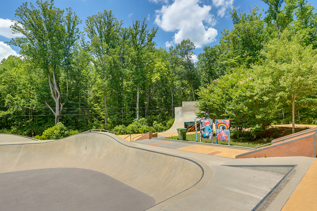 A sunny skate park with a large concrete bowl in the foreground and several ramps and railings off to the right.
