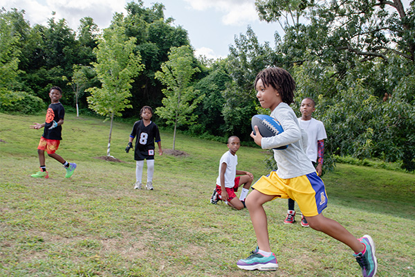 Five youths play football in a grassy field near some trees.