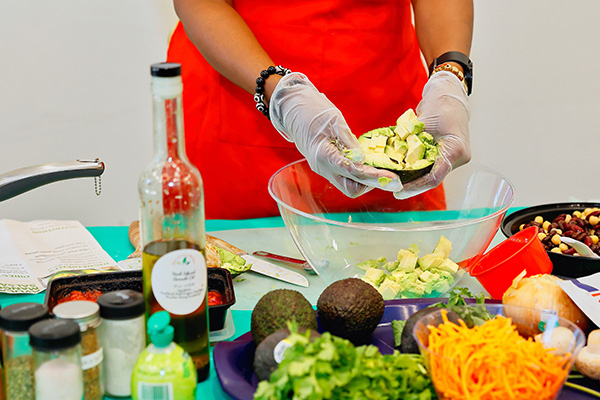 A person in a red apron cutting up an avocado and putting it into a bowl. They are surrounded by other ingredients across the table.