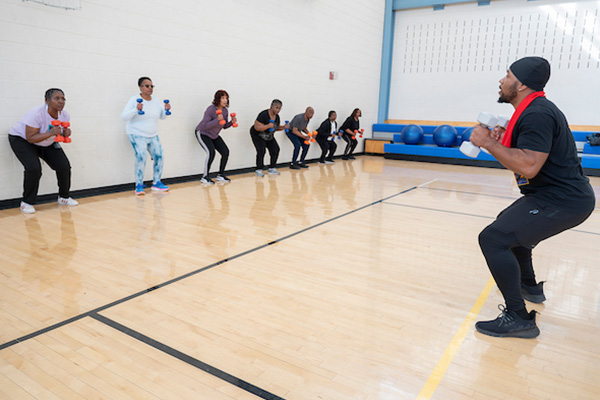 In a gymnasium, an instructor leads a class of seniors who are positioned along the wall, doing squats while holding hand weights in both hands.