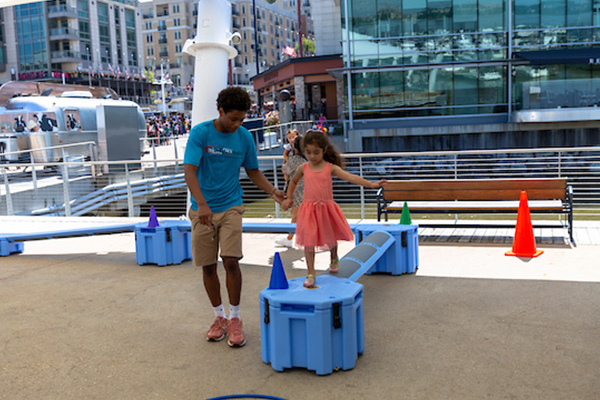A teen holds a child's hand while she walks along a blue balance beam-like structure on an outdoor obstacle course near a glass building.