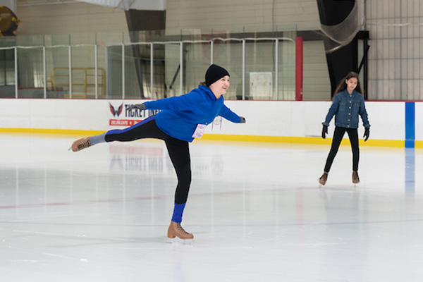 On an indoor ice rink, a woman in blue skates with one leg extended out behind her, and her arms out, smiling.