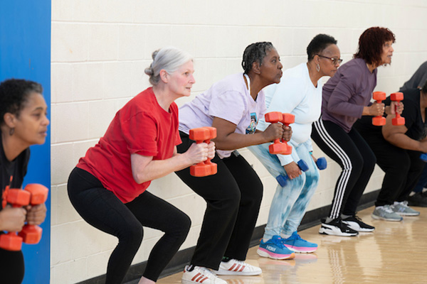 In a gymnasium, six senior women in exercise clothes perform a squat while holding weights in their hands.