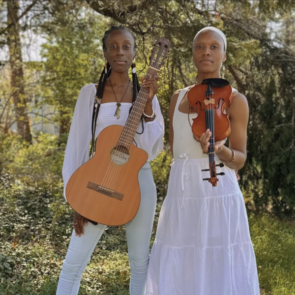 Two women standing outdoors in white garments holding string instruments, one with a guitar and one with a violin.