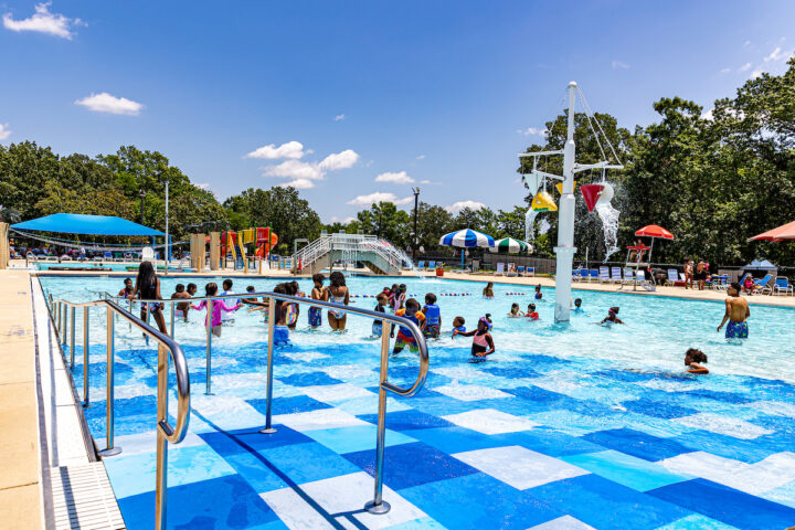 People enjoying a large outdoor pool with large ramp and a central splash feature. Behind this pool are additional splash park elements.