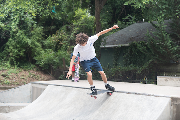 A skater riding down a ramp at an outdoor park. Trees fill the background behind the ramp with a young child skater watching.