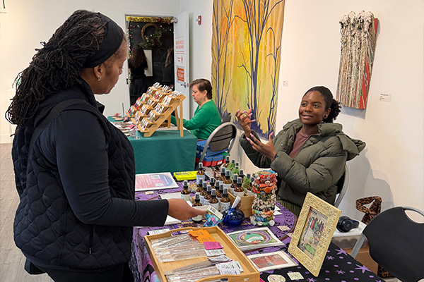 A customer talks with a vendor at a craft fair, where several long tables with colorful tablecloths display artisan wares.