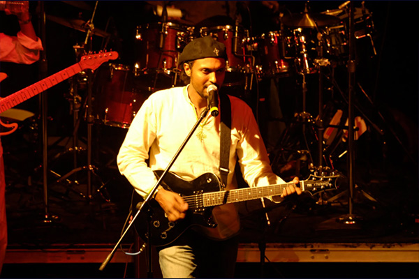 A man sings into a microphone while playing a black guitar. A drum kit and a bass guitar player are visible behind him.