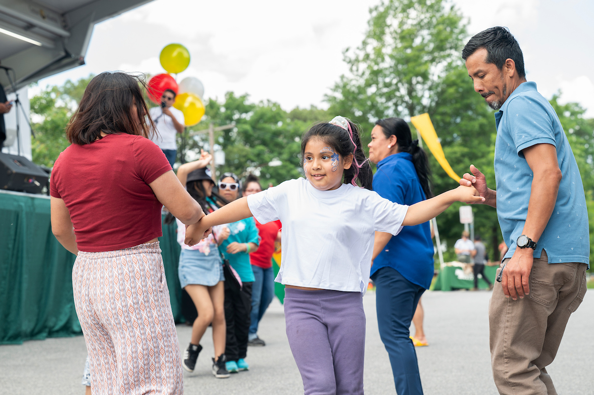 Child with face paint holds hands while dancing with adults at an outdoor community event near a stage.