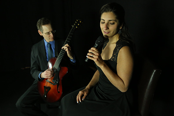 A seated woman sings into a microphone next to a seated man playing classical guitar.