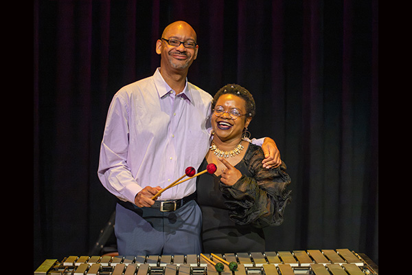 A smiling man and woman stand behind a large xylophone. The man holds mallets in one hand and has his other arm around the woman's shoulders.