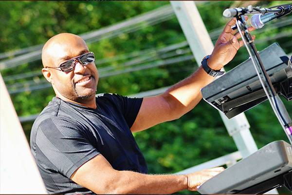 A smiling man wearing sunglasses plays electronic keyboards in the sunshine.