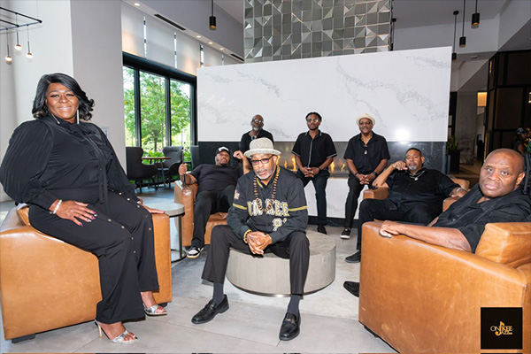 Eight adults in black clothing pose on armchairs and tables in a lobby.