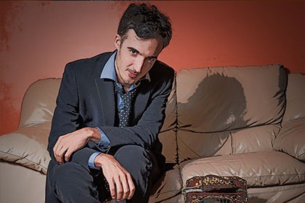 A man in a dark suit sits on a brown leather couch.