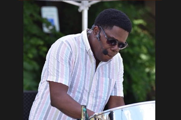 A man wearing sunglasses plays a steel drum under a tent outside.