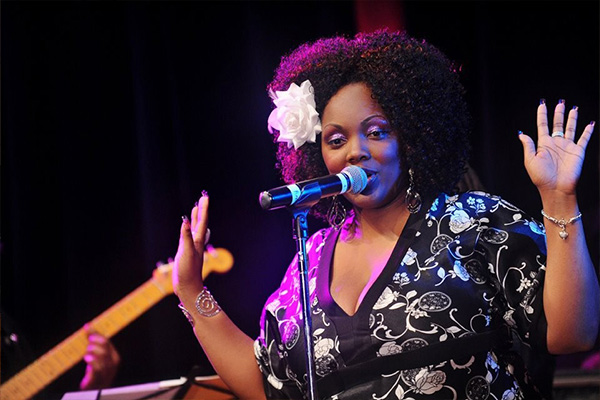 A woman in a black-and-white top with a flower in her hair sings into a microphone on a stage. A guitarist is visible behind her.