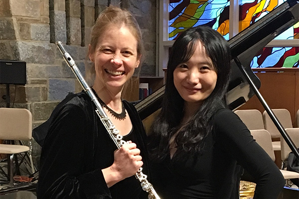 Two smiling women in black pose next to a grand piano. One holds a silver flute.